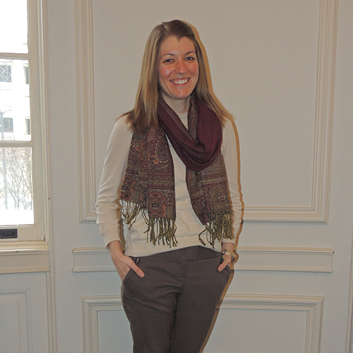 Heather Atkinson Caucasian female wearing a white long sleeve sweater and violet scarf with her hands in the pockets of her gray slacks.