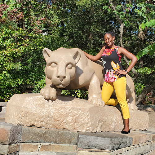 Shana Clarke African American woman with yellow pants and flowery top standing by Penn State Nittany Lion Shrine.