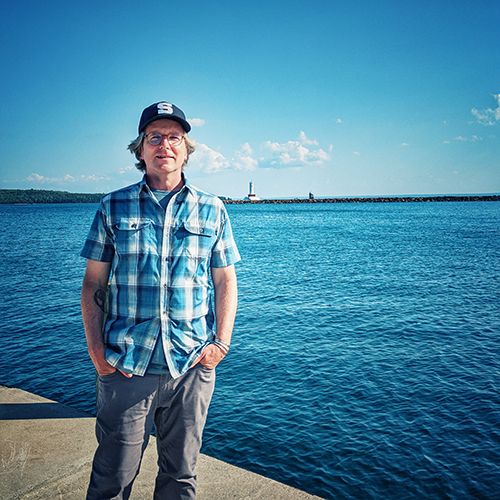 David Smith Middle-aged Caucasian male wearing jeans and a blue plaid shirt standing with his hands in his pockets in front of a lake.