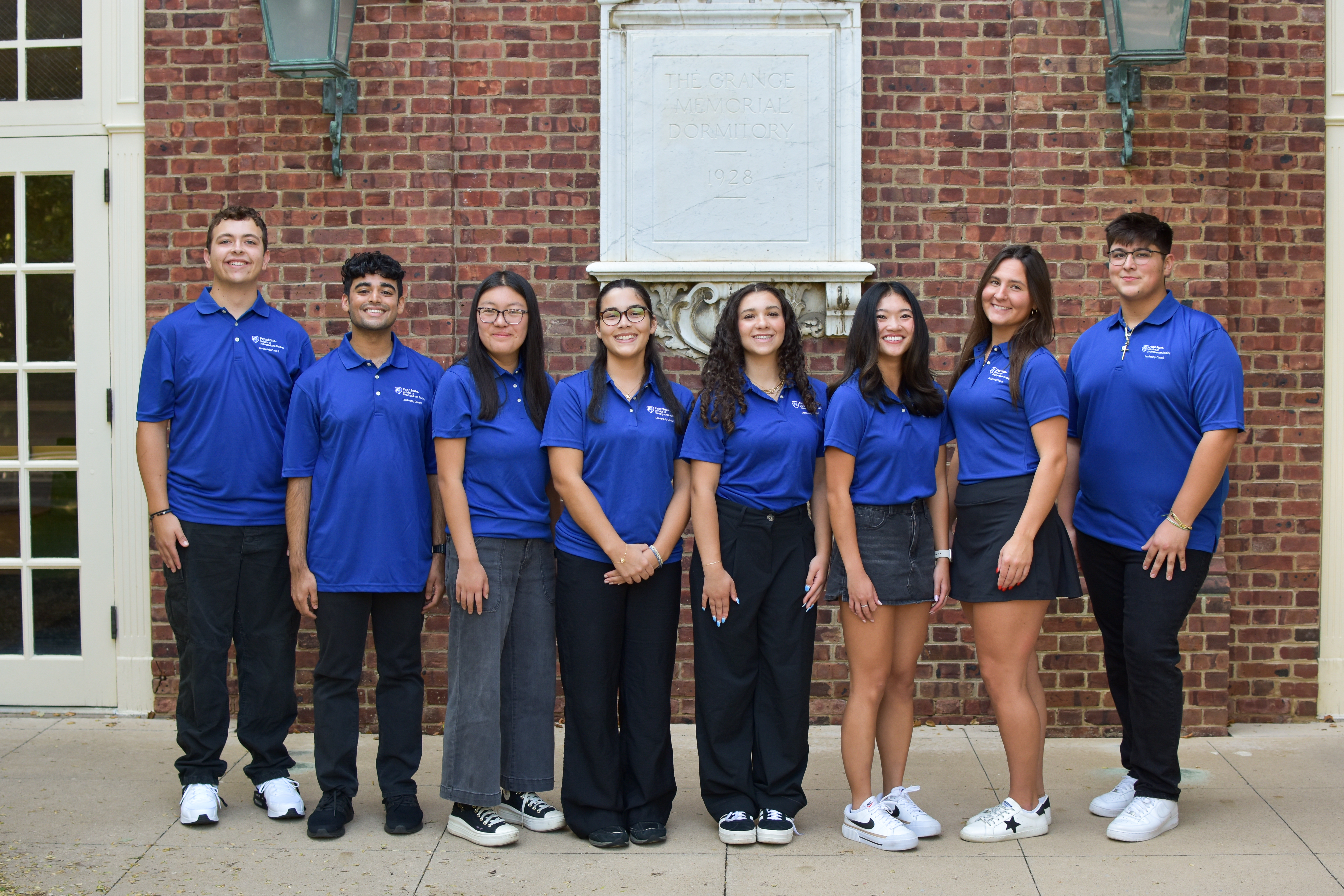Eight student leaders in blue polos stand smiling in front of The Grange Memorial Dormitory, showcasing a welcoming presence.